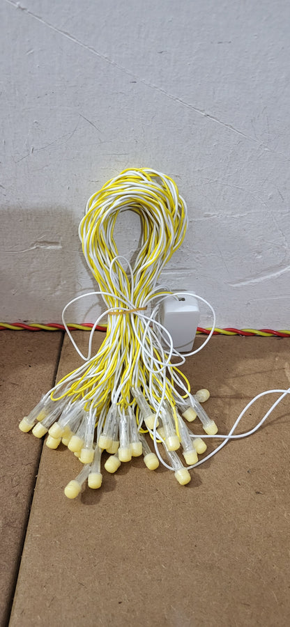 A bundle of miniature string lights with yellow and white wires and small warm-white LED bulbs, tied together with a white cordless light adapter, resting on a cardboard box against a concrete wall.